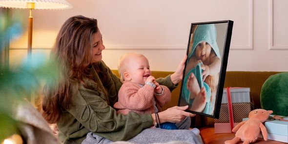 Frau mit einem Baby auf dem Schoß in einem Wohnzimmer, die personalisierte Wandkunst mit dem Baby als Motiv hält, während das Baby sie ansieht und kichert, passend zu posterXXLs Geschenkideen mit Fotos zu Babys erstem Weihnachten.