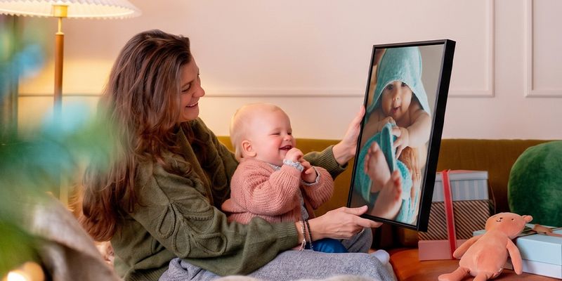 Frau mit einem Baby auf dem Schoß in einem Wohnzimmer, die personalisierte Wandkunst mit dem Baby als Motiv hält, während das Baby sie ansieht und kichert, passend zu posterXXLs Geschenkideen mit Fotos zu Babys erstem Weihnachten.