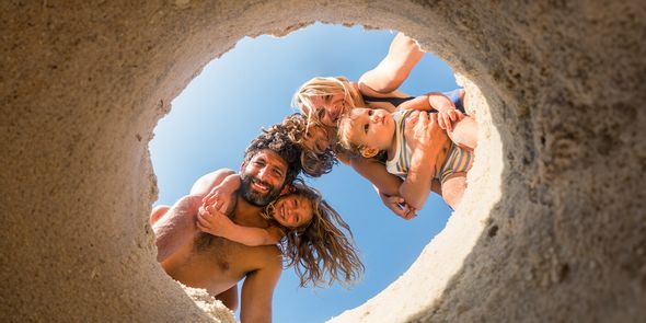 Eine vierköpfige Familie am Strand – Mutter, Vater, Tochter und kleiner Sohn – schaut in ein Loch im Sand und fängt damit einen verspielten Sommermoment ein, der zu Strandfotos und Strandfotografie inspiriert.