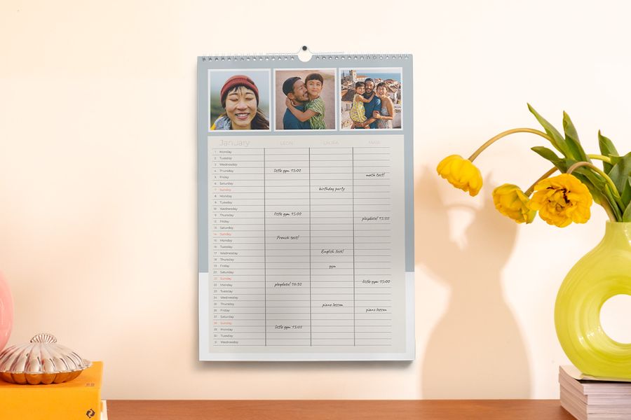 Ein Familienplaner hängt an der Wand neben einer Vase und einigen Büchern, und veranschaulicht posterXXLs Tipp, mit einem personalisierten Planer während des Schuljahres organisiert zu bleiben.