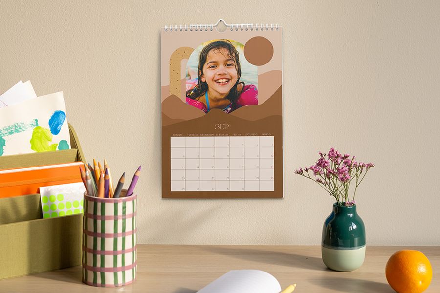 Ein personalisierter Kalender, der den September und ein Foto eines jungen Mädchens zeigt, hängt über einem Schreibtisch mit einem Stiftebecher, einer Vase mit Blumen und einem Organizer mit Bastel- und Schulmaterial, und veranschaulicht posterXXLs Tipp, einen fokussierten Lernbereich für das Schuljahr zu schaffen.
