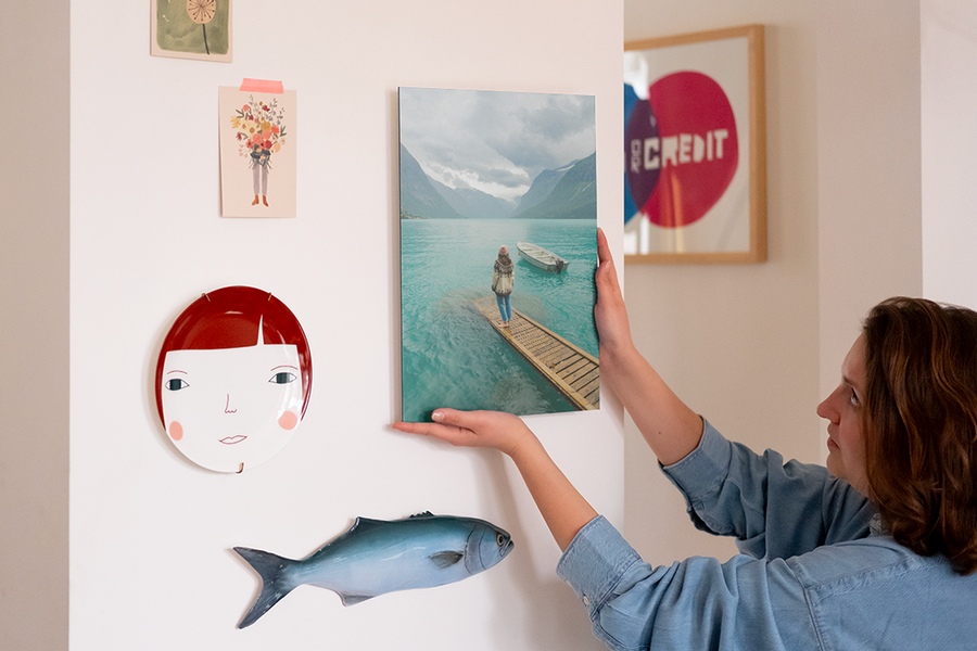 Eine Frau hängt personalisierte Wandkunst an ihre Wand, neben einem dekorativen Teller und einigen Fotoabzügen, und veranschaulicht posterXXLs letzten Schritt, die Wandkunst aufzuhängen und den Look zu vollenden.