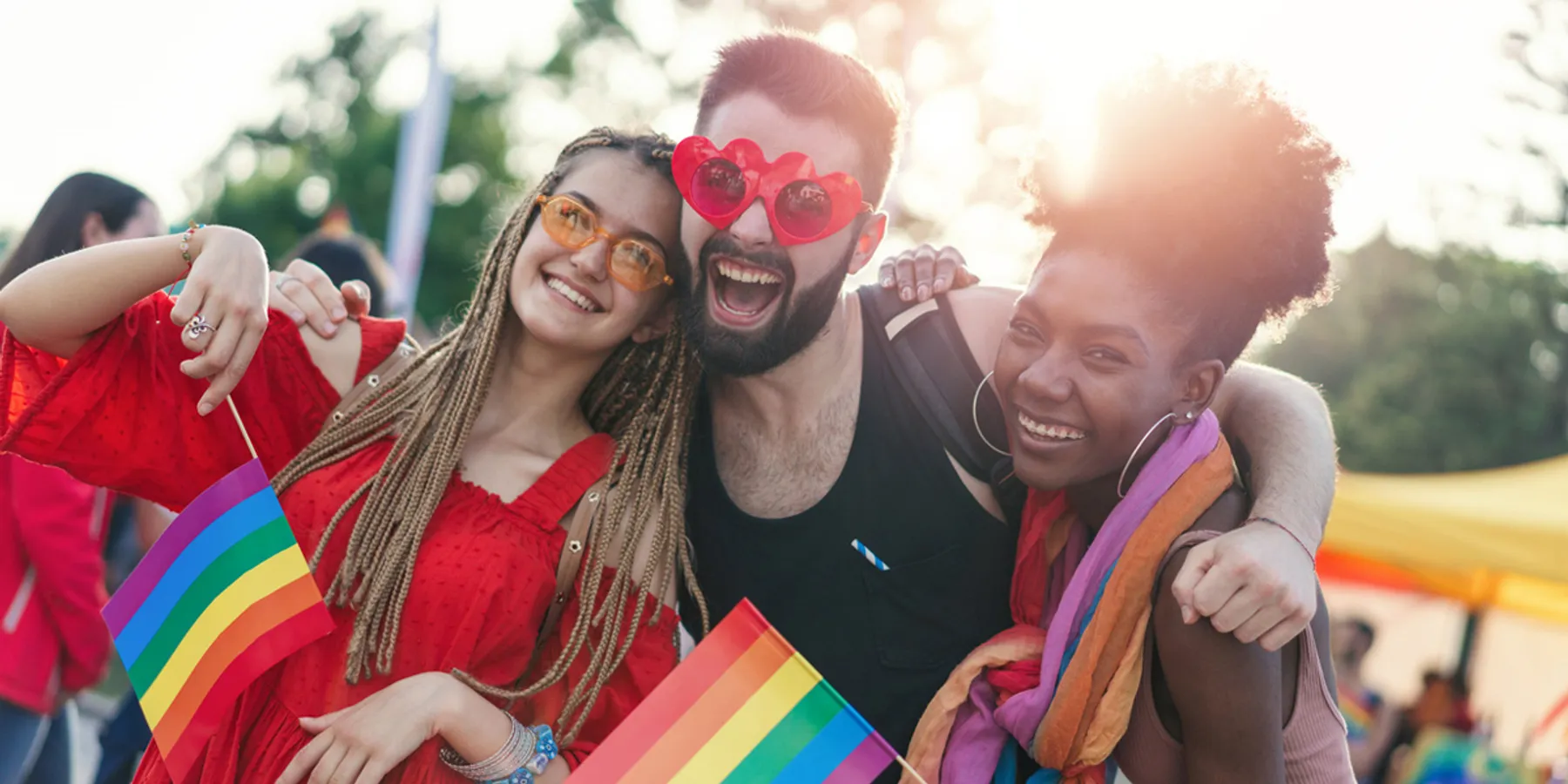 Drei Freunde lächeln bei einer Pride-Veranstaltung, halten Regenbogenflaggen in der Hand und tragen Sonnenbrillen – als Ausdruck von Freude und Zusammenhalt der LGBTQI+ Pride.