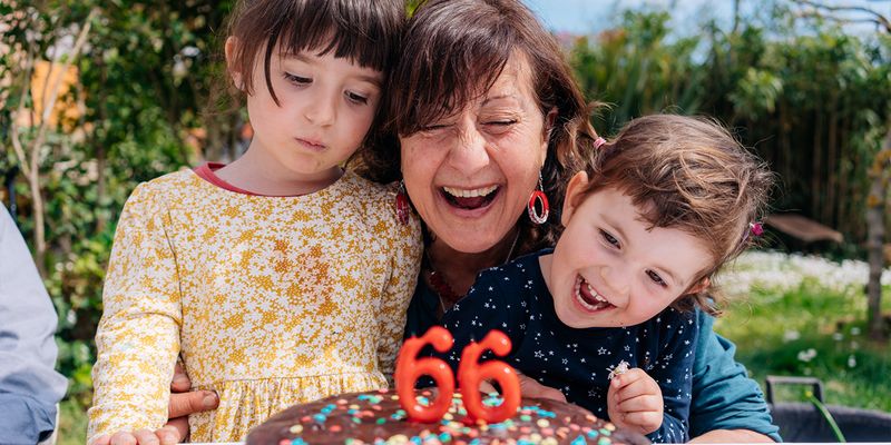 Eine Frau lächelt neben ihrer Geburtstagstorte mit Kerzen, die '66' zeigen, flankiert von einem kleinen Mädchen und einem kleinen Jungen, und stellt posterXXLs ultimativen Geburtstagsgeschenke-Ratgeber vor.