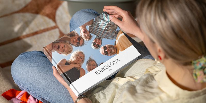 Une femme regarde un livre photo intitulé « Barcelone », avec une image de nombreux visages souriants en famille regardant vers l'appareil photo, illustrant le guide de Photobox pour créer un livre photo de vacances entre amis.