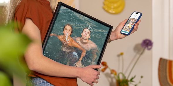Eine Frau hält ein gerahmtes Sommerfoto von einem Mann und einem kleinen Jungen, die im Wasser spielen, unter dem Arm, während sie auf ihrem Handy weitere Fotos auswählt, und veranschaulicht posterXXLs Ratgeber, Urlaubsfotos in Wandkunst zu verwandeln.