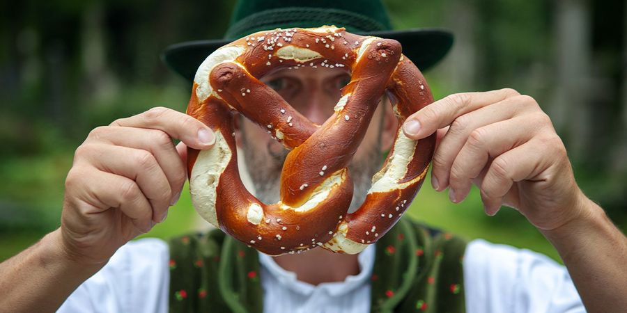 Ein Mann in traditioneller bayerischer Tracht hält eine große Brezel vor sein Gesicht, und stellt posterXXLs Ratgeber zu den besten Oktoberfest-Städten in Europa vor.