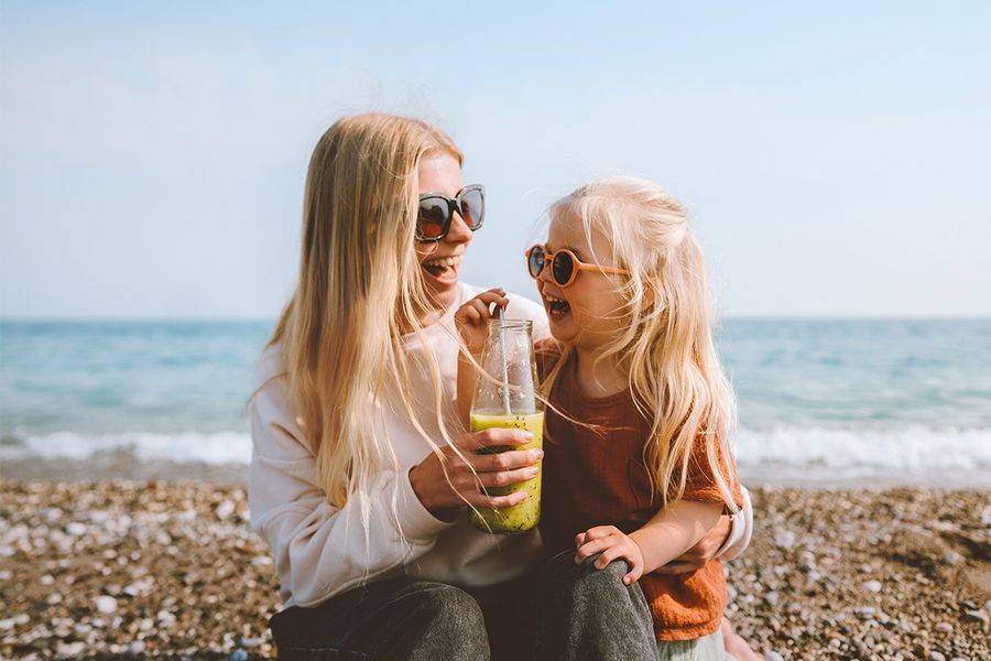 Mutter und Tochter lächeln am Strand, während sie einen Smoothie trinken.