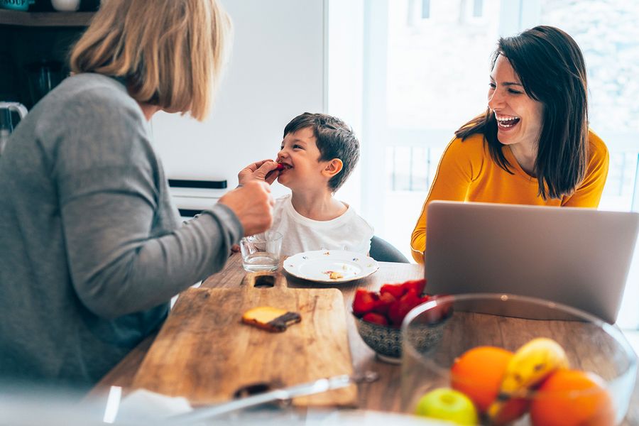Lächelnde Mehrgenerationenfamilie am Tisch; Großmutter füttert ihren Enkel, während die Mutter ein Notebook zur Erstellung eines Jahrbuchs nutzt.