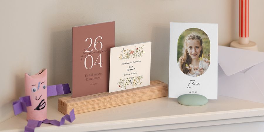 Eine Auswahl an verschiedenen Einladungen zur Erstkommunion von posterXXL, die gemeinsam in einem Regal präsentiert werden.