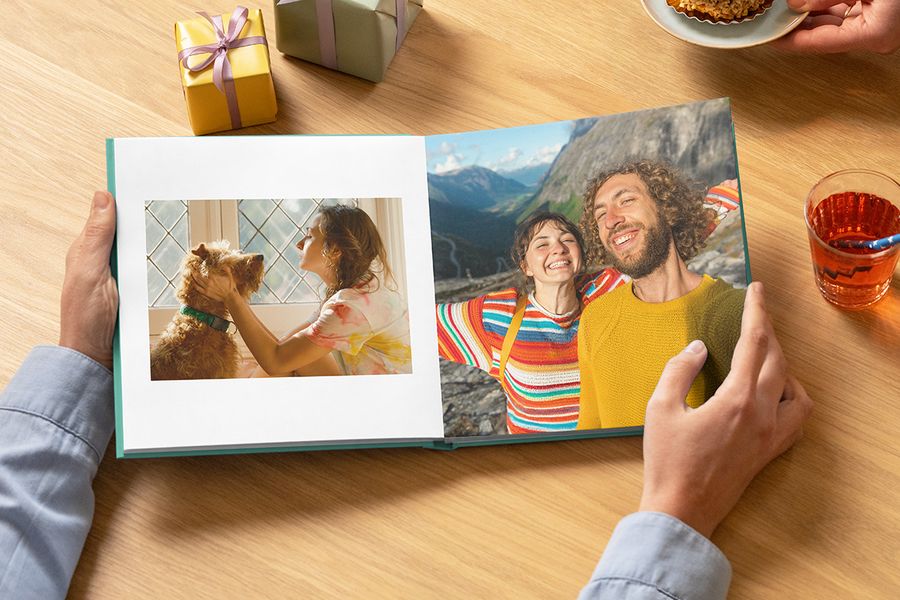 Ein offenes Hardcover-Fotobuch zum Geburtstag von posterXXL mit einer Frau und ihrem Hund sowie einem lächelnden Paar auf Reisen als Inspiration für ein Geburtstags fotobuch.