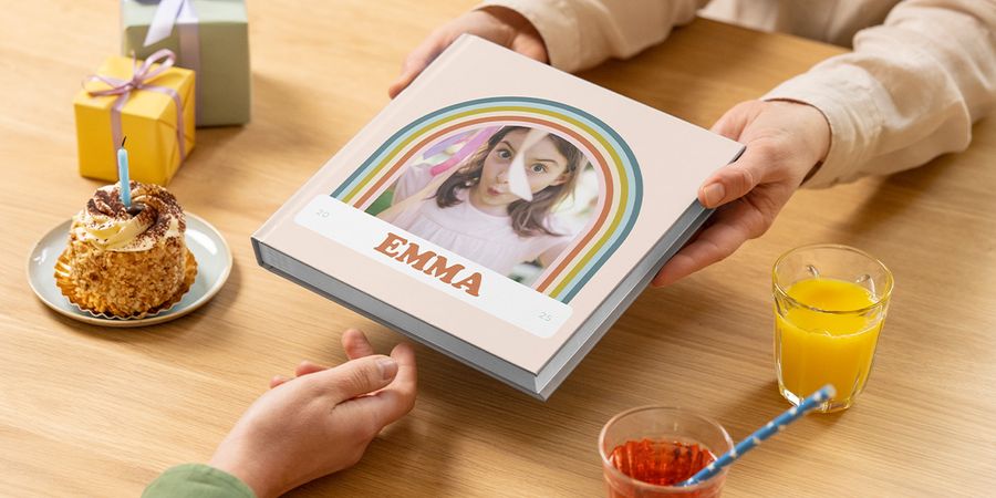 Ein Fotobuch zum Geburtstag für Emma wird über einen Tisch mit Kuchen und einem Geschenk gereicht; ein persönliches Fotobuch zum Geburtstag oder Geburtstags fotobuch von posterXXL.