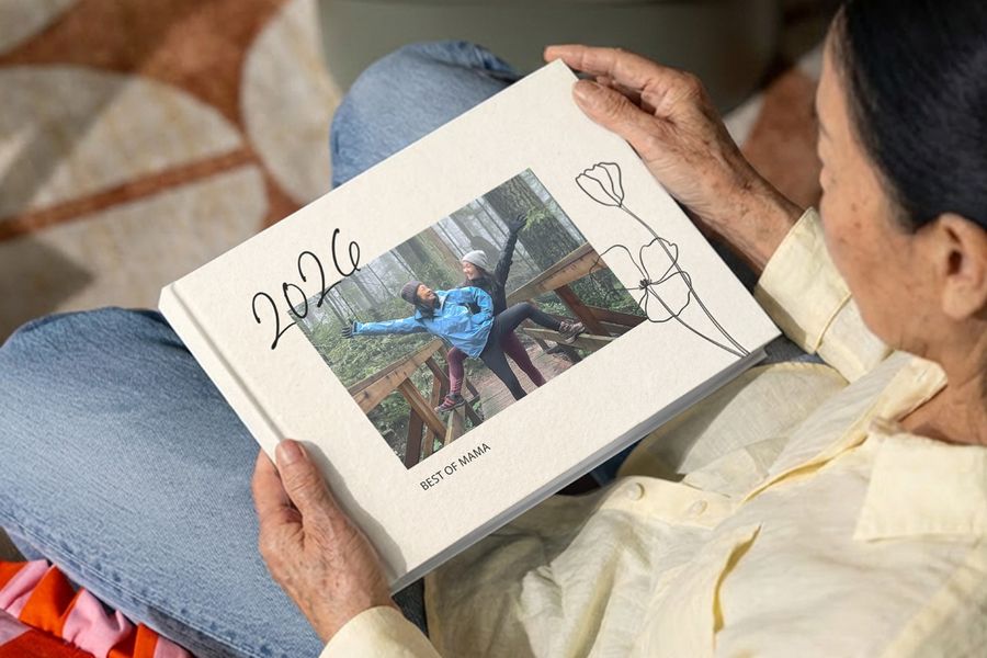 Eine Frau schaut sich ein Yearbook-Fotobuch von posterXXL mit dem Titel „Best of Mama“ an, auf dessen Cover sie selbst zusammen mit ihrer erwachsenen Tochter fröhlich auf einer Brücke zu sehen ist – als Inspiration für ein Fotobuch zum Muttertag und ein persönliches Muttertagsgeschenk.
