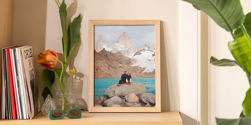 Ein gerahmtes Landschaftsfoto auf einem Holzregal neben Büchern und Blumen, ein Beispiel für Bilder aufhängen ohne bohren mit posterXXL.