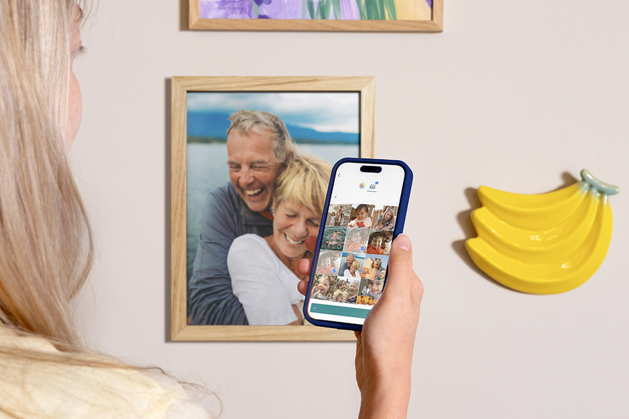 Eine Frau hält ihr Smartphone hoch, um zu zeigen, wie einfach es ist, posterxxl Fotoabzüge über die App zu bestellen, während im Hintergrund gerahmte Fotoabzüge an der Wand hängen – als Inspiration für Fotos online bestellen und Fotos vom Handy drucken.