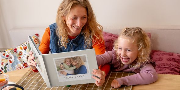 Eine Mutter und ihre kleine Tochter schauen gemeinsam ein personalisiertes posterXXL Fotobuch mit einem Foto von ihnen auf dem Cover an – als Inspiration für ein Fotobuch zum Muttertag und ein persönliches Muttertagsgeschenk.
