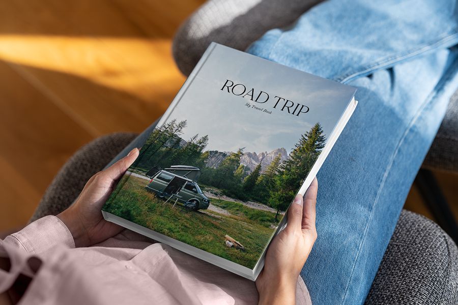 Ein posterXXL Coffee Table Fotobuch mit dem Titel Road Trip liegt auf dem Schoß einer Person, mit einer Waldlandschaft und einem Wohnmobil auf dem Cover – als Inspiration für ein Reise-Coffee-Table-Book.