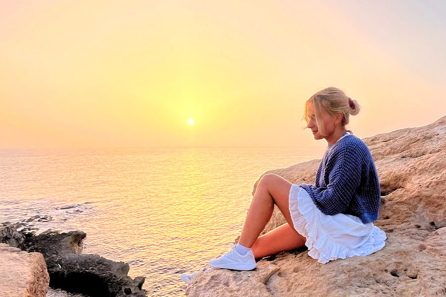 Eine Frau sitzt auf Felsen am Meer, während hinter ihr die Sonne untergeht – ein Beispiel dafür, wie das goldene Licht der Abendsonne warme und wunderschöne Fotos entstehen lässt.