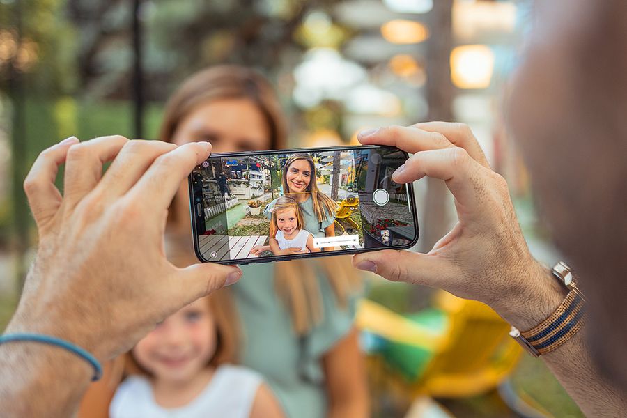 Eine Hand hält ein Smartphone, mit dem eine lächelnde Mutter und ihre Tochter im hellen Sonnenlicht fotografiert werden – als Beispiel dafür, wie man mit dem Handy bei starkem Sonnenlicht fotografiert.