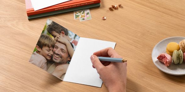 Eine geöffnete personalisierte posterXXL Vatertagskarte zeigt einen Vater mit seinen zwei Söhnen, daneben liegt ein Stift zum Schreiben einer Nachricht neben Büchern und einem Teller Macarons – als Inspiration dafür, was man in eine Vatertagskarte schreiben kann.