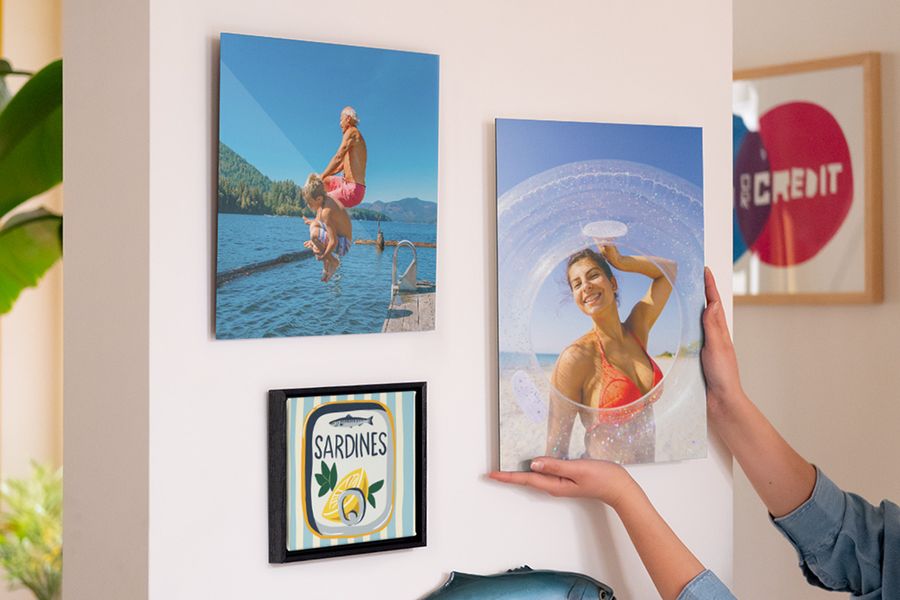 Eine Bilderwand mit personalisierter posterXXL Wanddeko zeigt eine lächelnde Frau am Strand, einen Mann und Jungen beim Sprung ins Meer sowie eine Sardinendosen-Illustration – als Inspiration für sommerliche Deko Ideen und sommerliche Wanddeko.