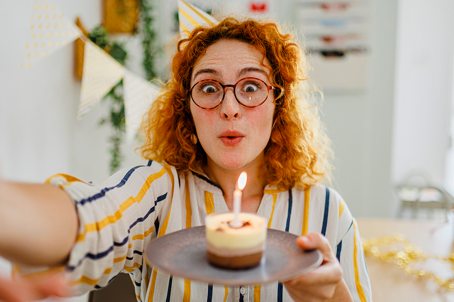 Eine Frau mit roten Locken hält einen Cupcake mit Kerze in der Hand und inspiriert damit zu Geburtstagswünschen für Kollegen und Geburtstagskarten von posterXXL.