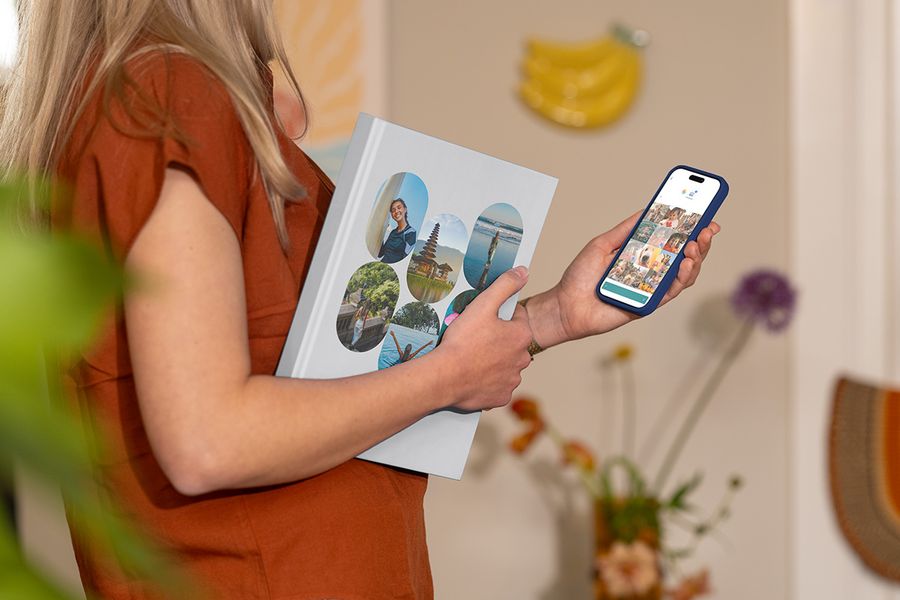 Eine Frau hält ein geschlossenes posterXXL Urlaubsfotoalbum mit Familienreisefotos auf dem Cover in der Hand und schaut gleichzeitig Reisebilder auf ihrem Smartphone an – als Illustration dafür, wie man die richtigen Fotos für ein personalisiertes Reise-Fotobuch auswählt.