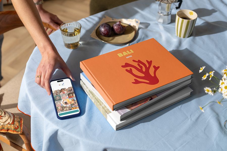 Ein Stapel verschiedener posterXXL Reise-Fotobücher liegt neben einer Hand, die ein Smartphone mit einem ausgewählten Urlaubsfoto hält, obenauf ein Fotobuch Bali 2026 mit Korallenmotiv auf dem Cover, und zeigt, wie man ein personalisiertes Reise-Fotobuch gestaltet.