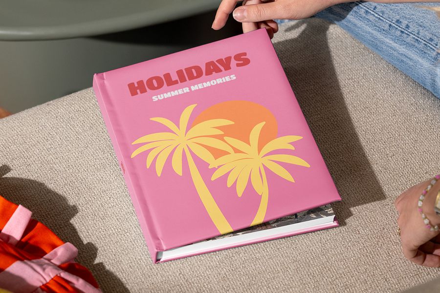 Ein geschlossenes posterXXL Reise-Fotobuch mit dem Titel „Holidays, summer memories“ und illustrierten Palmen auf einem rosa Cover wird von Händen gehalten, die es gleich öffnen wollen – als Inspiration dafür, wie man ein Urlaubsfotoalbum strukturiert und seine Reisegeschichte erzählt.