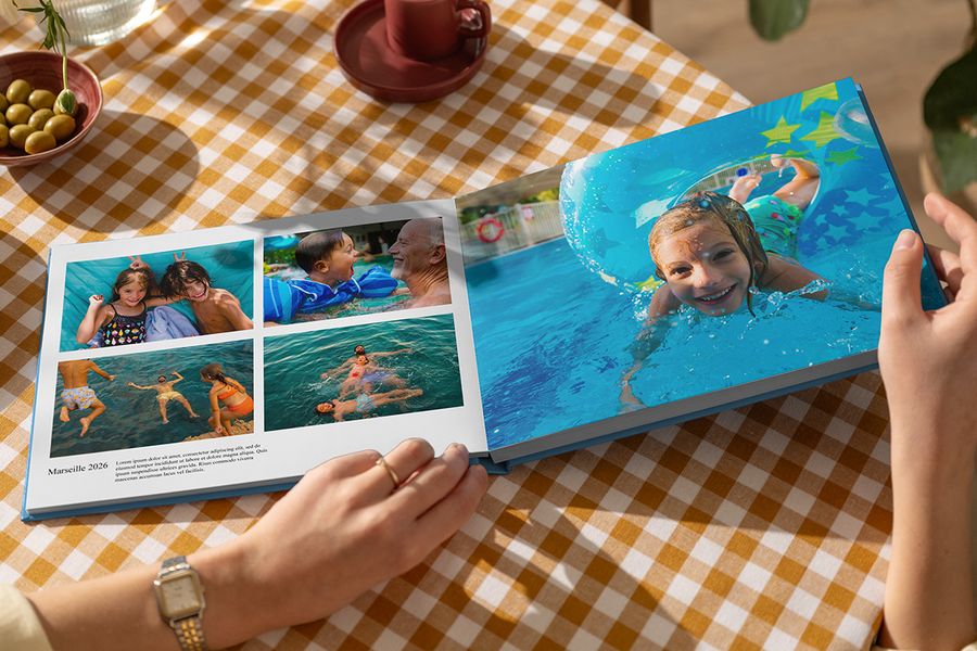 Ein geöffnetes posterXXL Urlaubsfotobuch zeigt sommerliche Familienfotos beim Spielen im Wasser, mit einer eindrucksvollen Doppelseite und einer weiteren Seite mit dem Titel Marseille 2026 samt Bildunterschrift und Beschreibung – als Illustration dafür, wie Texte ein personalisiertes Reise-Fotobuch lebendig machen.