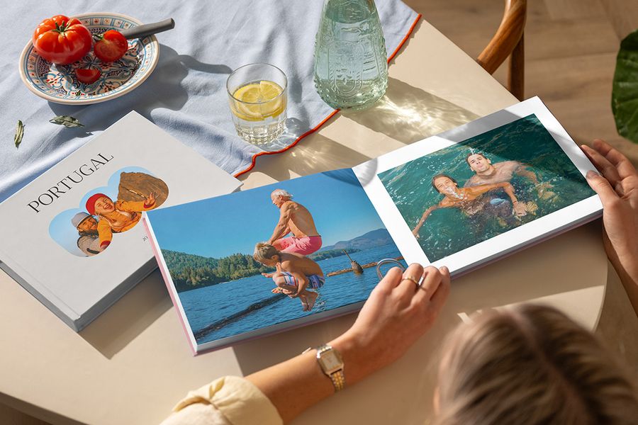Zwei verschiedene posterXXL Reise-Fotobücher sind zu sehen: Eine Frau blättert durch ein geöffnetes Urlaubsfotobuch mit Familienfotos am Meer, daneben liegt ein geschlossenes Portugal-Fotobuch – ein personalisiertes Reise-Fotoalbum voller Urlaubserinnerungen.