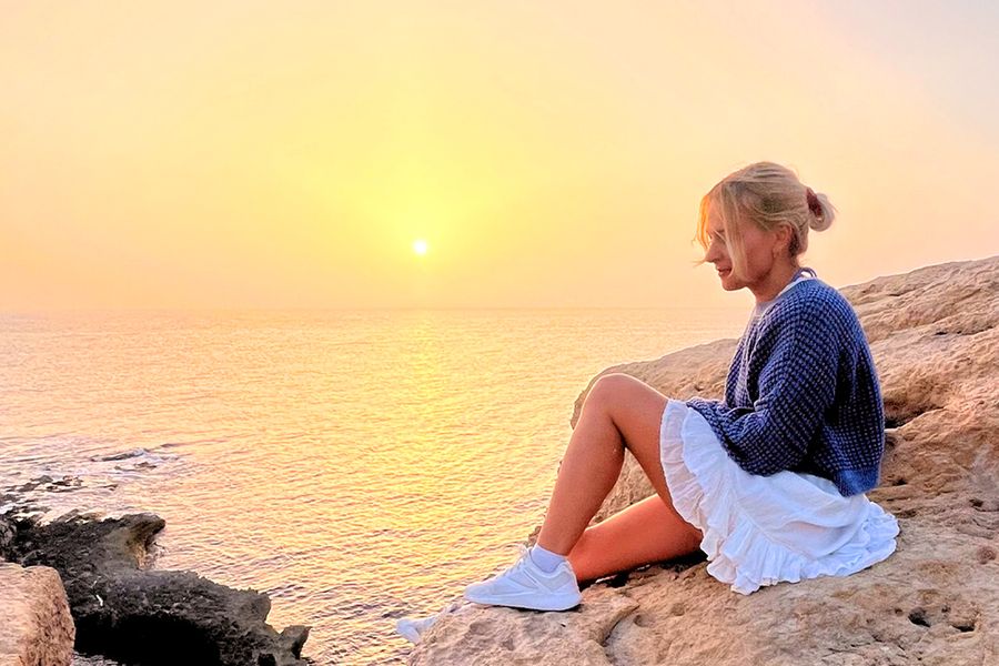 Eine Frau sitzt auf Steinen und blickt hinaus auf das Meer und den Sonnenuntergang, was den Geist von posterXXLs abenteuerlichen Staycation-Ideen einfängt.