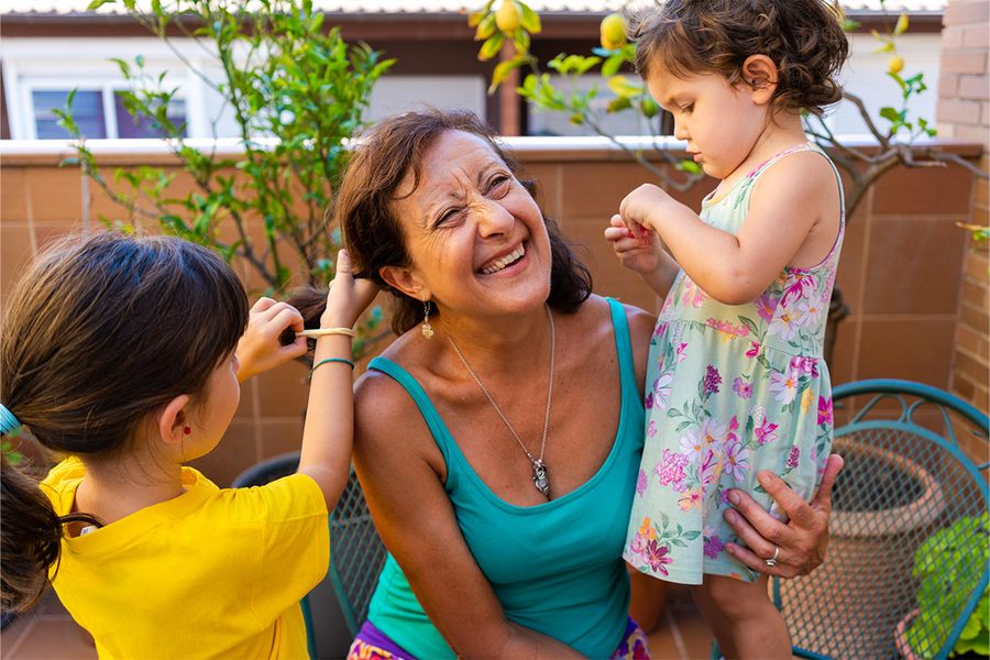 Eine Frau sitzt auf einem Balkon, während zwei junge Mädchen mit ihrem Haar spielen und es stylen, ein verspielter Moment aus posterXXLs Staycation-Ideen für Familien.