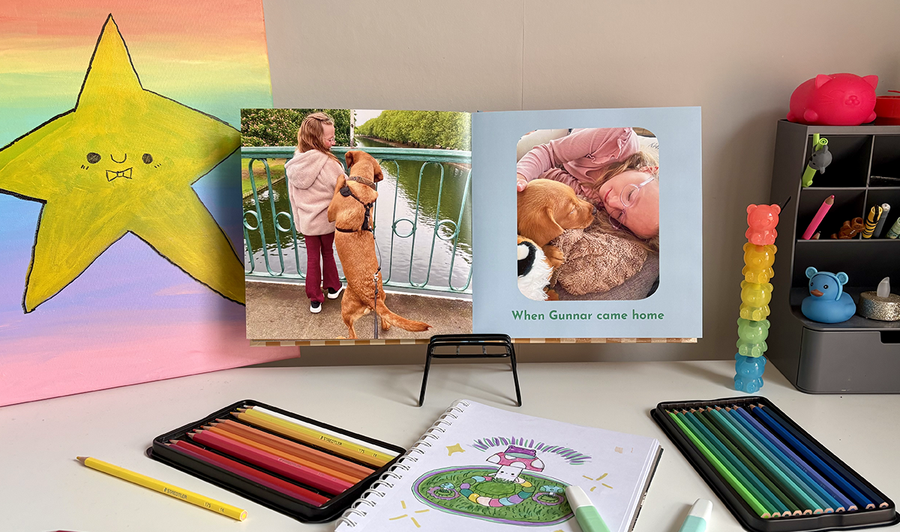 Ein aufgeschlagenes Kinderfotobuch liegt auf dem Schreibtisch eines Kindes, umgeben von Buntstiften und Kunstwerken, mit einem Foto eines Mädchens und ihres Hundes auf der linken Seite und einem weiteren Bild mit Bildunterschrift auf der rechten Seite, und veranschaulicht posterXXLs Tipps zum Mischen von Fotos und Kunstwerken in einem Familienfotobuch.