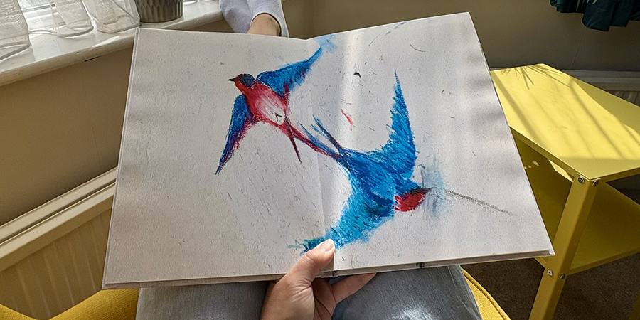 Ein aufgeschlagenes Kinderfotobuch mit einer doppelseitigen Vogelzeichnung liegt auf dem Schoß einer Frau, die in einem Sessel sitzt, und veranschaulicht posterXXLs Schritte zur Erstellung eines Familienfotobuchs aus den Zeichnungen eines Kindes.