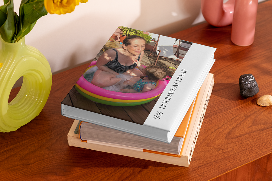 Ein Stapel Fotobücher zeigt oben ein Buch mit einer Frau und einem kleinen Kind in einem Planschbecken, mit dem Titel 'Holidays at Home', und veranschaulicht posterXXLs Fotobuch Titel Ideen für eine Staycation.