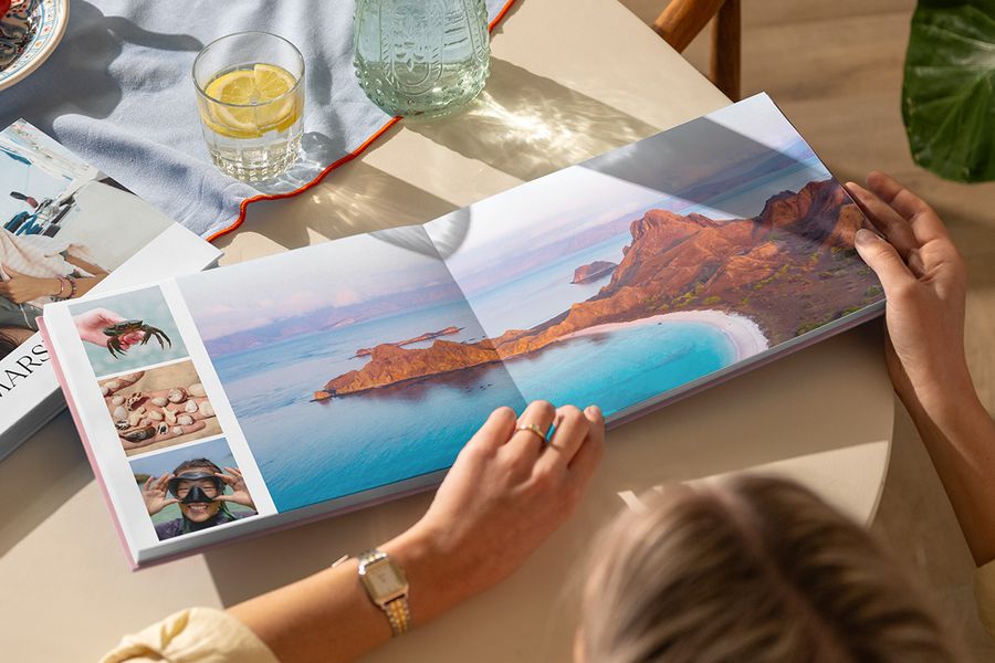Ein aufgeschlagenes Reisefotobuch auf einem Tisch zeigt eine Doppelseite mit einer wunderschönen Strandlandschaft neben drei Sommerfotos, und veranschaulicht posterXXLs Travel Chapter-Design aus der neuen Summer Travel Series.