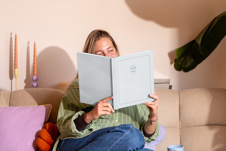 Eine Frau sitzt auf dem Sofa neben einem bunten Kissen und schaut sich ein personalisiertes Reisefotobuch mit dem Titel 'Oslo' an, das posterXXLs neue Summer Travel Series Designs zeigt.