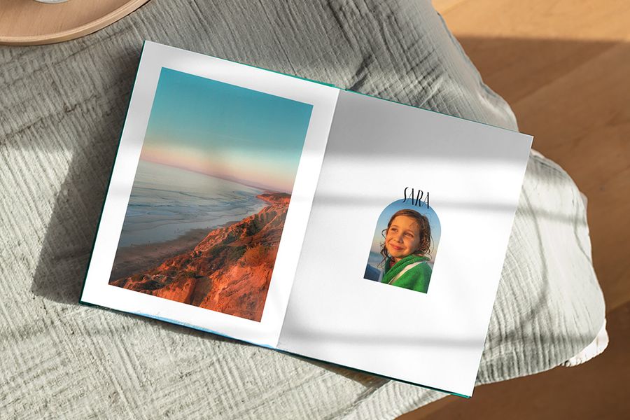 Ein aufgeschlagenes Fotobuch zeigt auf einer Seite eine wunderschöne Landschaft und auf der anderen ein Bild eines Mädchens mit der Bildunterschrift 'Sara', und veranschaulicht posterXXLs Artisan Masking-Design mit spielerischen Rahmen, inspiriert von Vintage-Postkarten und Briefmarken.