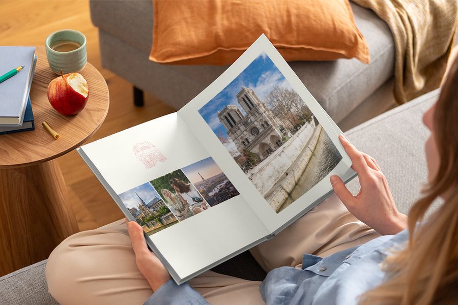 Eine Frau hält ein aufgeschlagenes Fotobuch mit verschiedenen Reisefotos auf ihrem Schoß, und veranschaulicht posterXXLs Graphic Getaways-Design, inspiriert von Vintage-Reiseplakaten und illustrierten Stadtplänen.