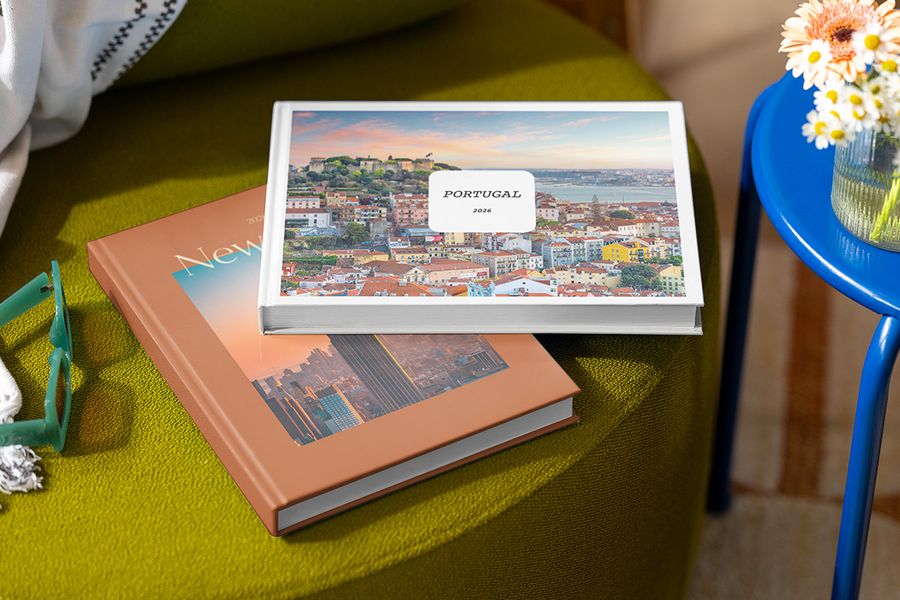 Zwei personalisierte Fotobücher liegen gestapelt auf einem Hocker, das obere mit dem Titel 'Portugal' und einem Bild von Portugal, darunter ein Buch mit dem Titel 'New York' und einem Bild der Stadt, und zeigen posterXXLs personalisierbare Summer Travel Series Designs.