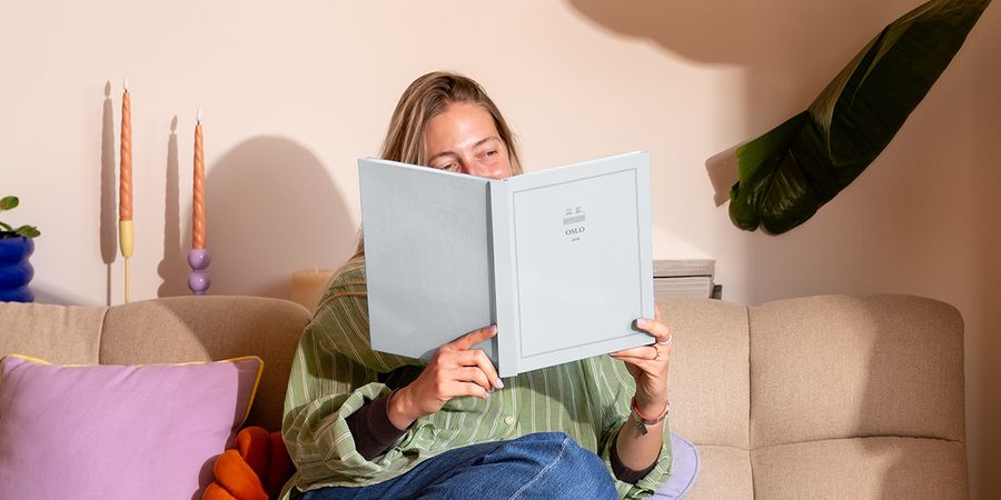 Eine Frau sitzt auf dem Sofa neben einem bunten Kissen und schaut sich ein personalisiertes Reisefotobuch mit dem Titel 'Oslo' an, das posterXXLs neue Summer Travel Series Designs zeigt.
