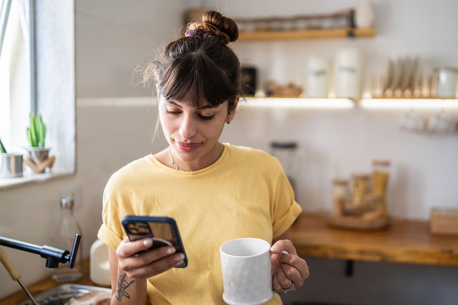 Eine Frau in ihrer Küche hält eine Tasse, während sie ihr Handy durchsieht, und veranschaulicht posterXXLs Tipps, um Foto-Ordnungsgewohnheiten aufzubauen, die wirklich bleiben.