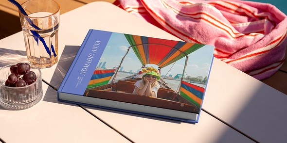 Ein großes querformatiges Reisefotobuch mit dem Titel 'Nomadic Anna' liegt auf einem Tisch, mit einem Foto eines Mädchens auf Reisen auf dem Cover, und veranschaulicht posterXXLs Fotobuch Titel Ideen für Reisen.