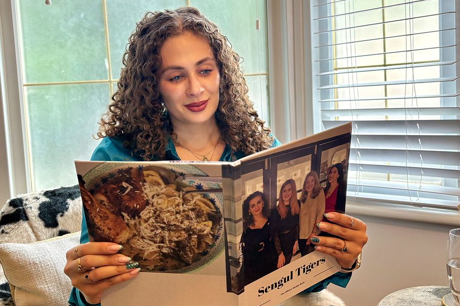 Sarah, die bei posterXXL arbeitet, sitzt in einem Stuhl mit Kuhfellmuster und betrachtet ihr persönliches Familienkochbuch, dessen Cover vier Frauen zeigt, darunter Sarah selbst, und veranschaulicht, wie ein Rezept-Fotobuch Familienrezepte und Erinnerungen zusammenbringt.