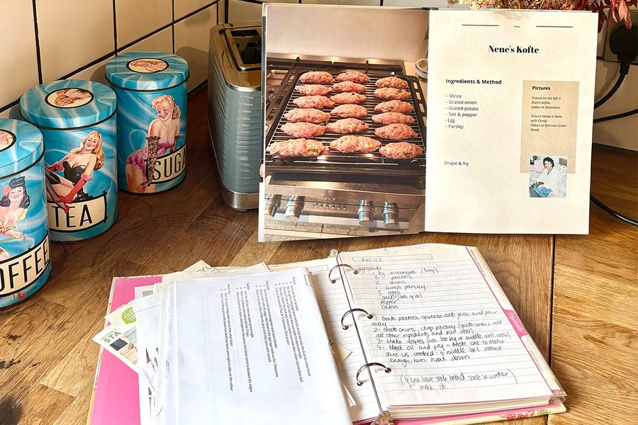 Sarahs aufgeschlagenes persönliches Kochbuch zeigt ein Rezept für Nenes Kofte, platziert vor einem alten Ordner, der handgeschriebene Familienrezepte zeigt, beide liegen auf einer Küchenarbeitsplatte, und veranschaulicht, wie sie Generationen von Rezepten in einem Buch zusammengeführt hat.
