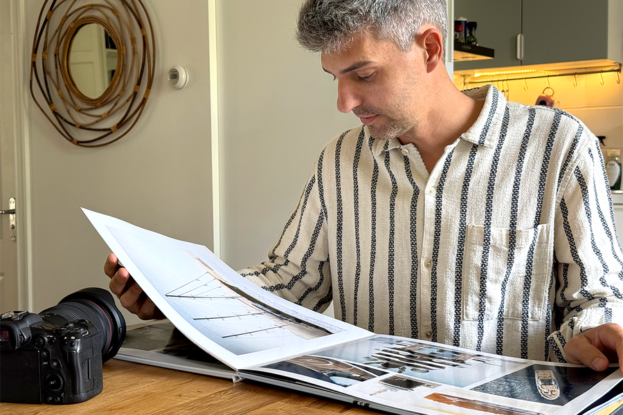 Charl van Rooy sitzt in seiner Küche und schaut auf sein aufgeschlagenes Portfolio, mit seiner Kamera neben sich auf dem Tisch, und veranschaulicht posterXXLs Ratgeber zum Erstellen eines Fotografie-Portfolios als Fotobuch.