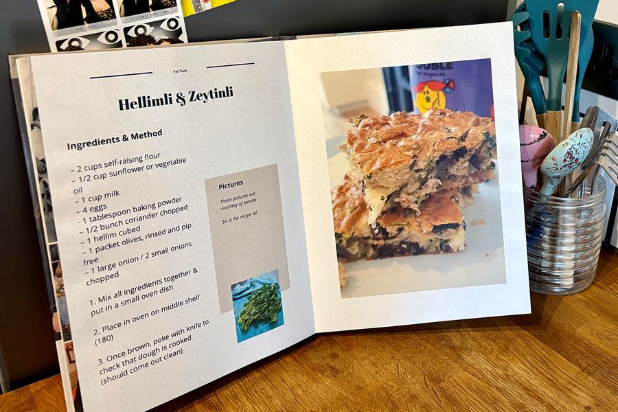Ein aufgeschlagenes persönliches Kochbuch liegt in einer Küche, mit einem Glas voller Löffel und Küchenutensilien im Hintergrund, und veranschaulicht posterXXLs Tipps zur Auswahl der Rezepte, bevor man sein eigenes Kochbuch erstellt.