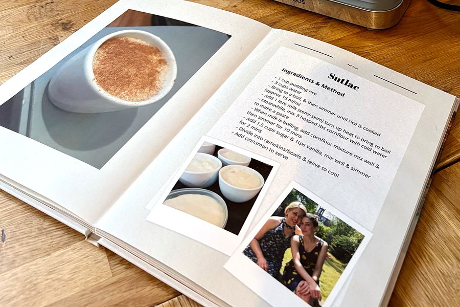 Sarahs aufgeschlagenes Rezeptbuch liegt auf der Küchenarbeitsplatte und zeigt sein Layflat-Fotobuchformat, mit Bildern von türkischem Reispudding auf der linken Seite und dem dazugehörigen Familienrezept auf der rechten Seite, und veranschaulicht posterXXLs Tipp, ein Layflat-Design zu wählen, das sich leicht zum Kochen nutzen lässt.