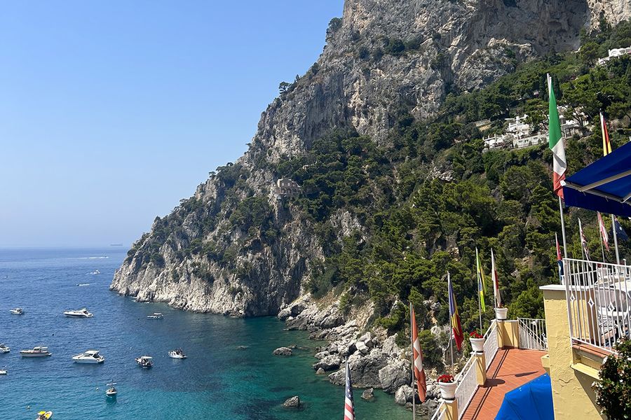 Eine Mittelmeerküste mit Hügeln an einem blauen, mit Booten gesprenkelten Meer, gesehen von einem Balkon im Vordergrund, und fängt den Charme eines September-Urlaubs in Europa ein.